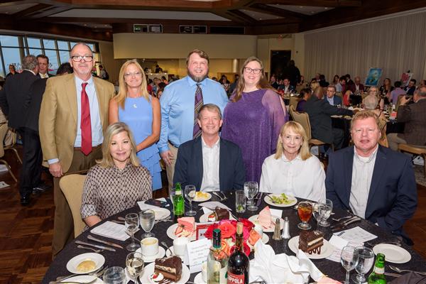 A group of eight well-dressed adults pose for a photo at a formal dinner event. Four individuals are standing behind a round table while four others are seated at the table. The table is set with glasses, plates of cake, salads, rolls, and bottles of wine and water. The background shows a large room